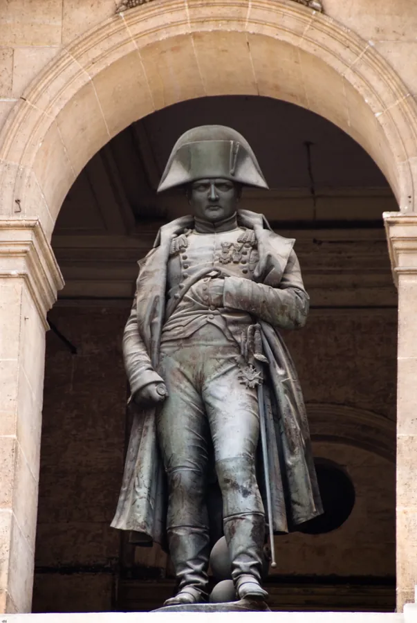 Photographie d’archive de la version précédente : statue de bronze de Napoléon debout, en redingote et bicorne, des boulets à ses pieds.