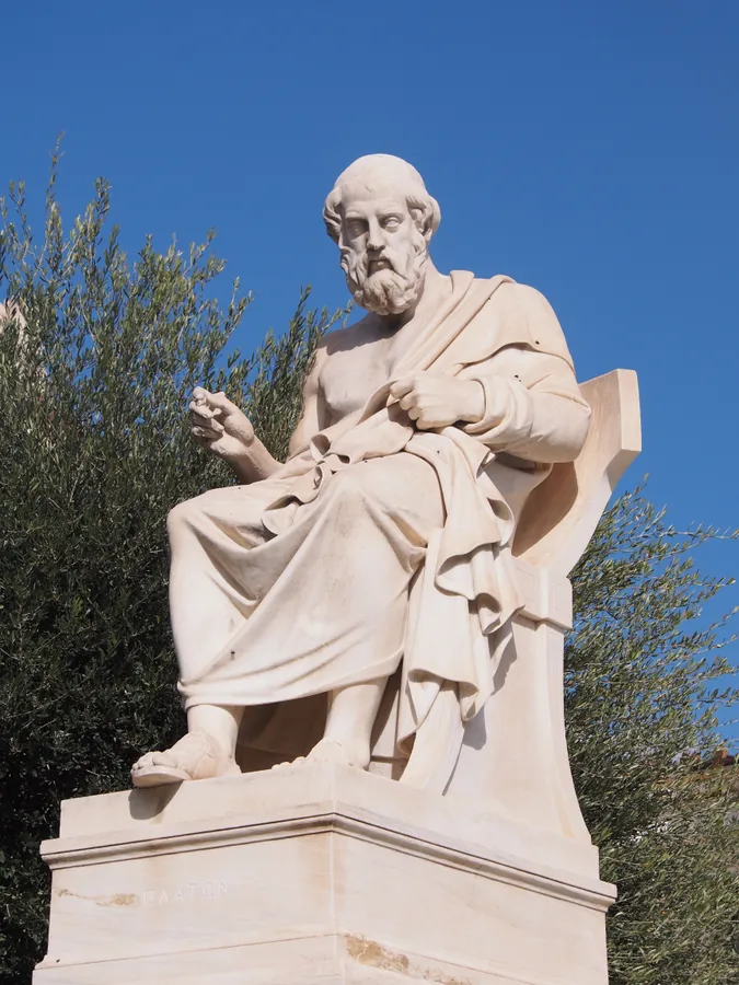 Photographie d’archive de la version précédente : statue de marbre de Platon assis, barbu, drapé, devant l’Académie d’Athènes.