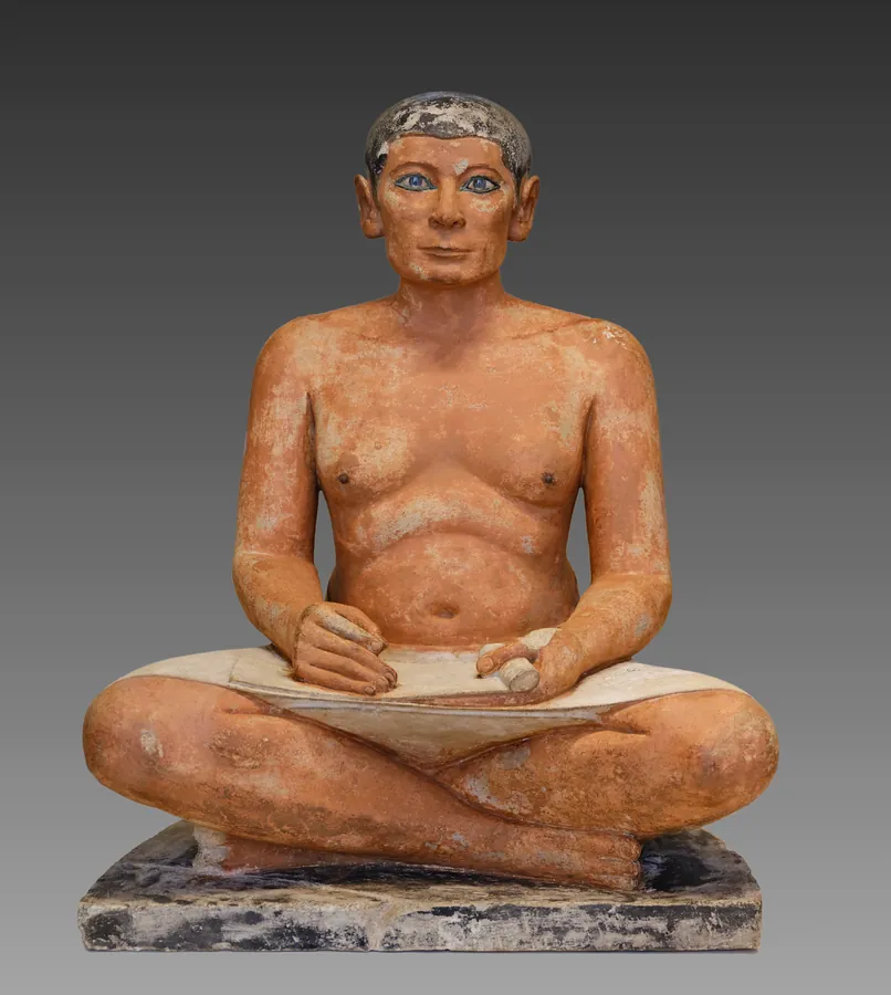 Photographie d’archive de la version précédente : statue de calcaire peint d’un scribe assis en tailleur, un papyrus déroulé sur les genoux, le regard fixe.